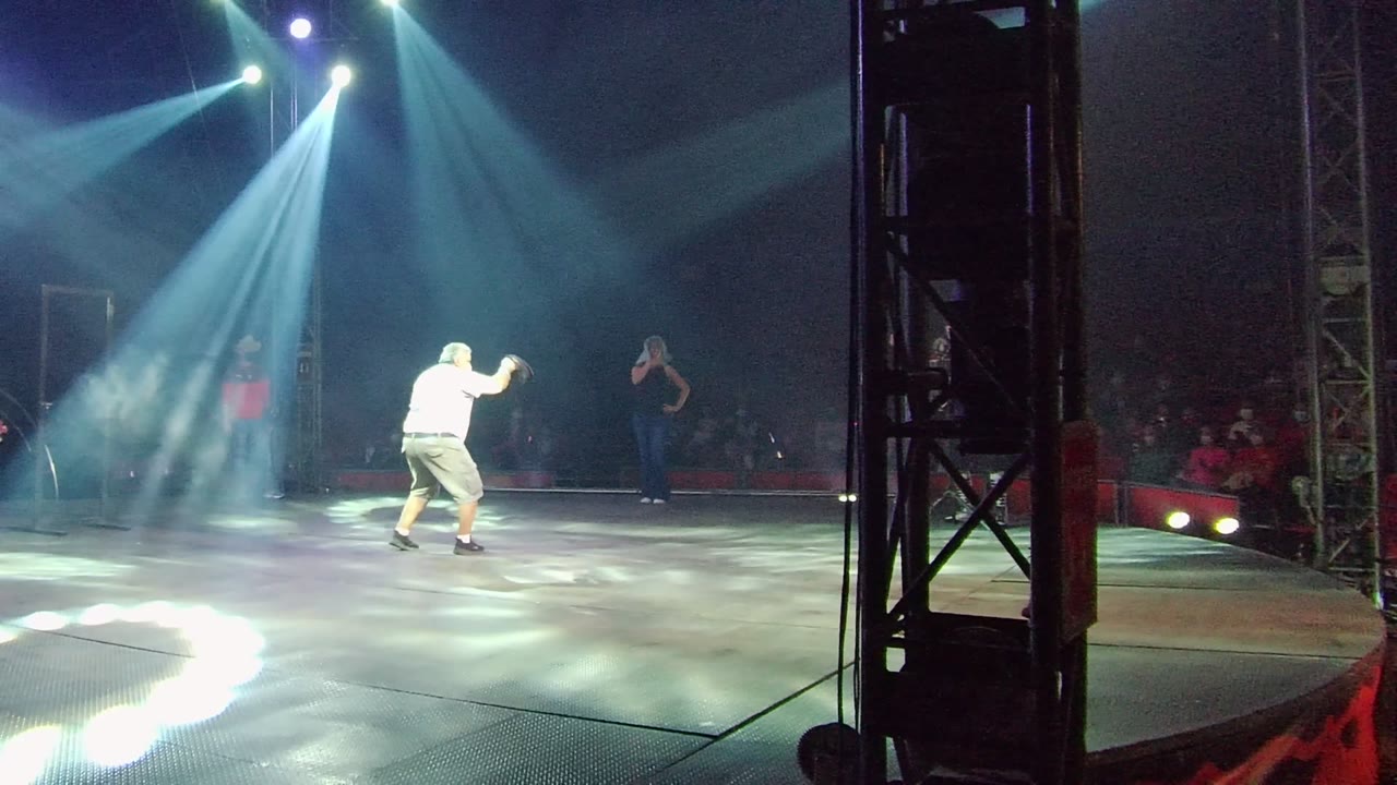 In San Jose del Cabo, Mexico, the dancer raises one arm while standing in the lit midground, with a dark vertical truss at right and audience seating behind the stage.