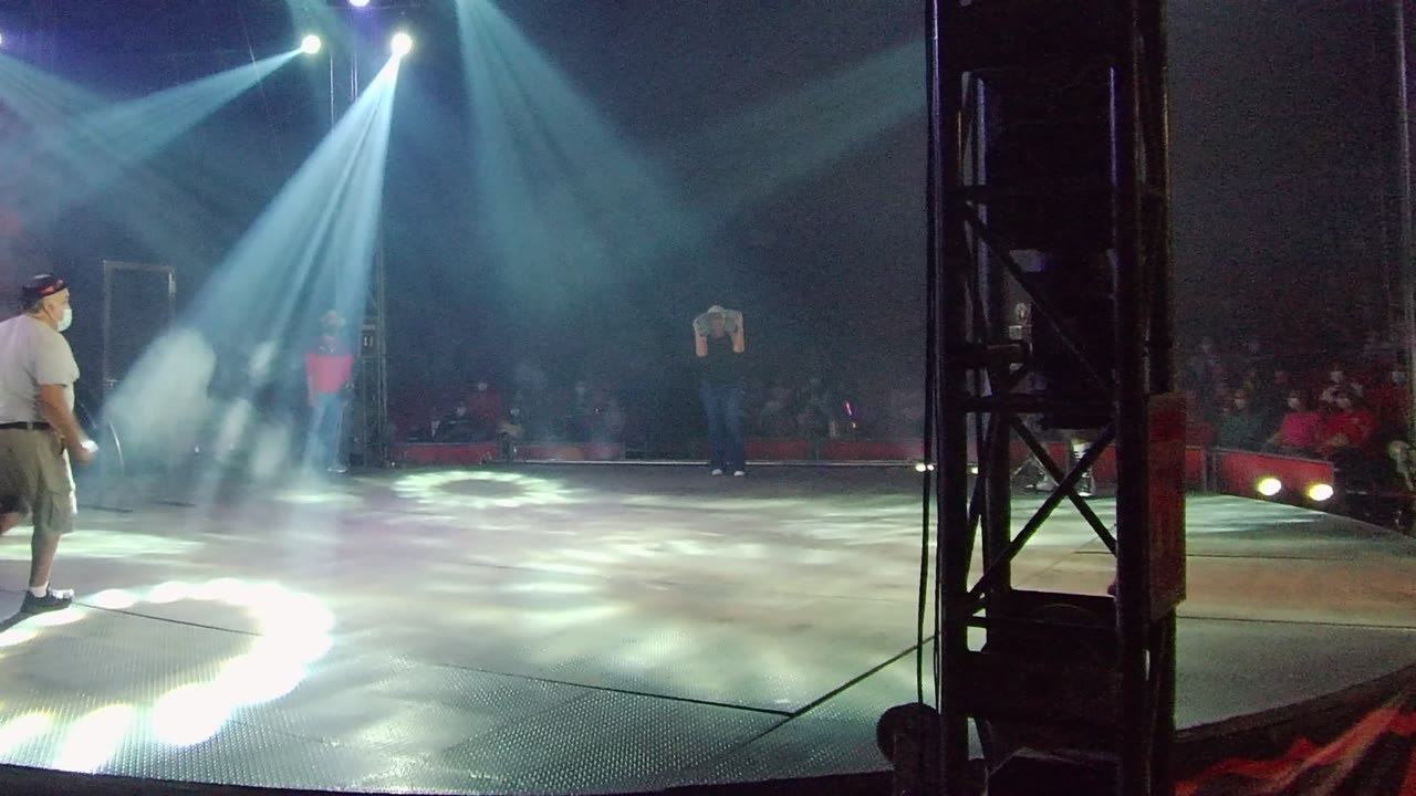 A performer in a white shirt stands near the left edge of a smoky stage in San Jose del Cabo, Mexico, with blue-white spotlights and a dark audience behind a red barrier.