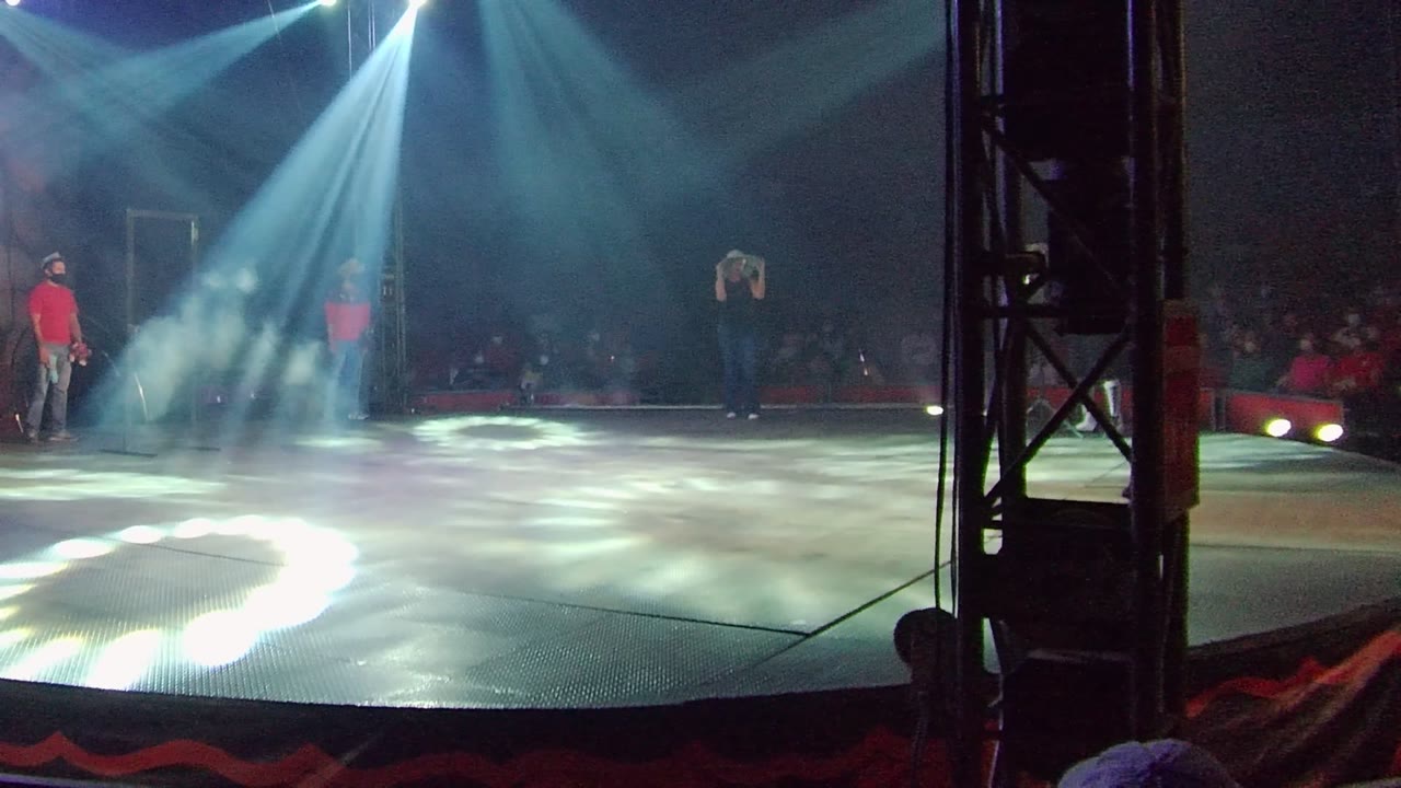 At 0:02, overhead white beams and stage haze dominate the frame while two performers remain separated across the stage and the audience sits in shadow behind them.