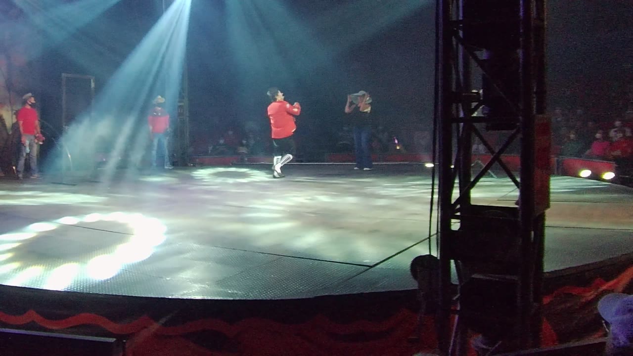 A red-shirt dancer stands on a spotlighted stage in San Jose del Cabo, Mexico, with haze, floor light patterns, and audience members visible behind the performance area.