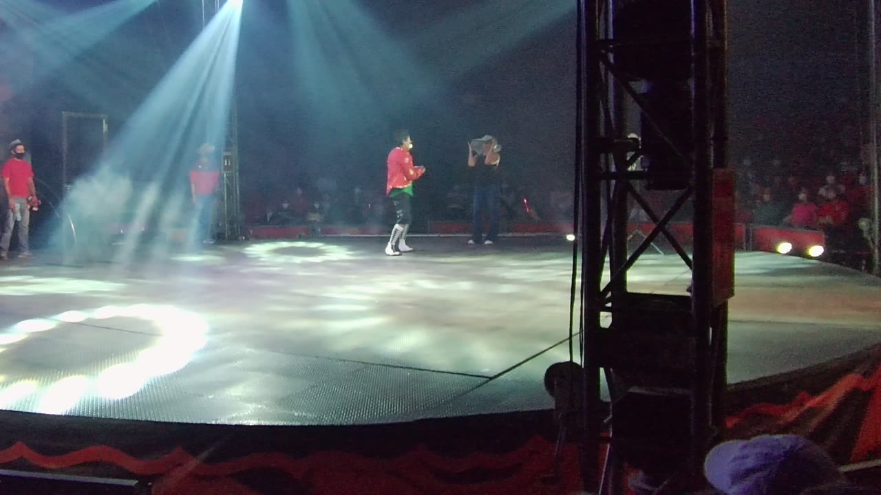 A red-shirt performer moves across the stage in San Jose del Cabo, Mexico, with pale spotlight patterns on the floor, haze in the air, and audience seating visible beyond the stage.