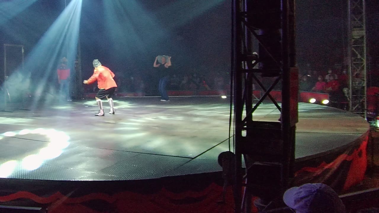 A dancer in an orange shirt stands left of center on a dark stage in San Jose del Cabo, Mexico, with white-blue spotlights overhead and audience members visible behind the performance area.