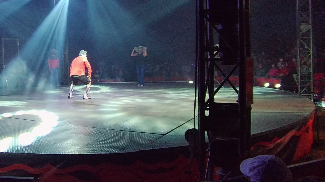 The performer bends low on the San Jose del Cabo stage, framed by a foreground truss at right, with white spotlight patterns on the floor and audience faces dimly visible in the background.