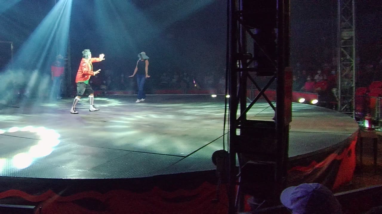 A performer in an orange shirt gestures across a smoky stage in San Jose del Cabo, Mexico, while another person stands farther back near the dark audience area.