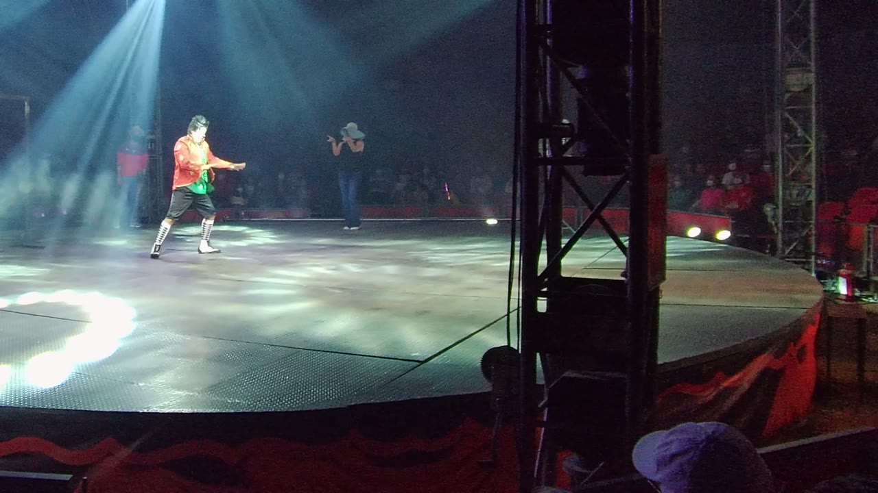 The lead performer steps across the lit circular stage in San Jose del Cabo, Mexico, with the second performer partly visible near the back and spectators in shadow.
