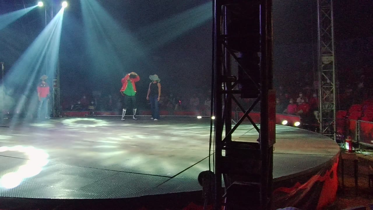 Three performers stand on a San Jose del Cabo stage as white spotlights spread across the floor and red audience seating sits in the dark background.