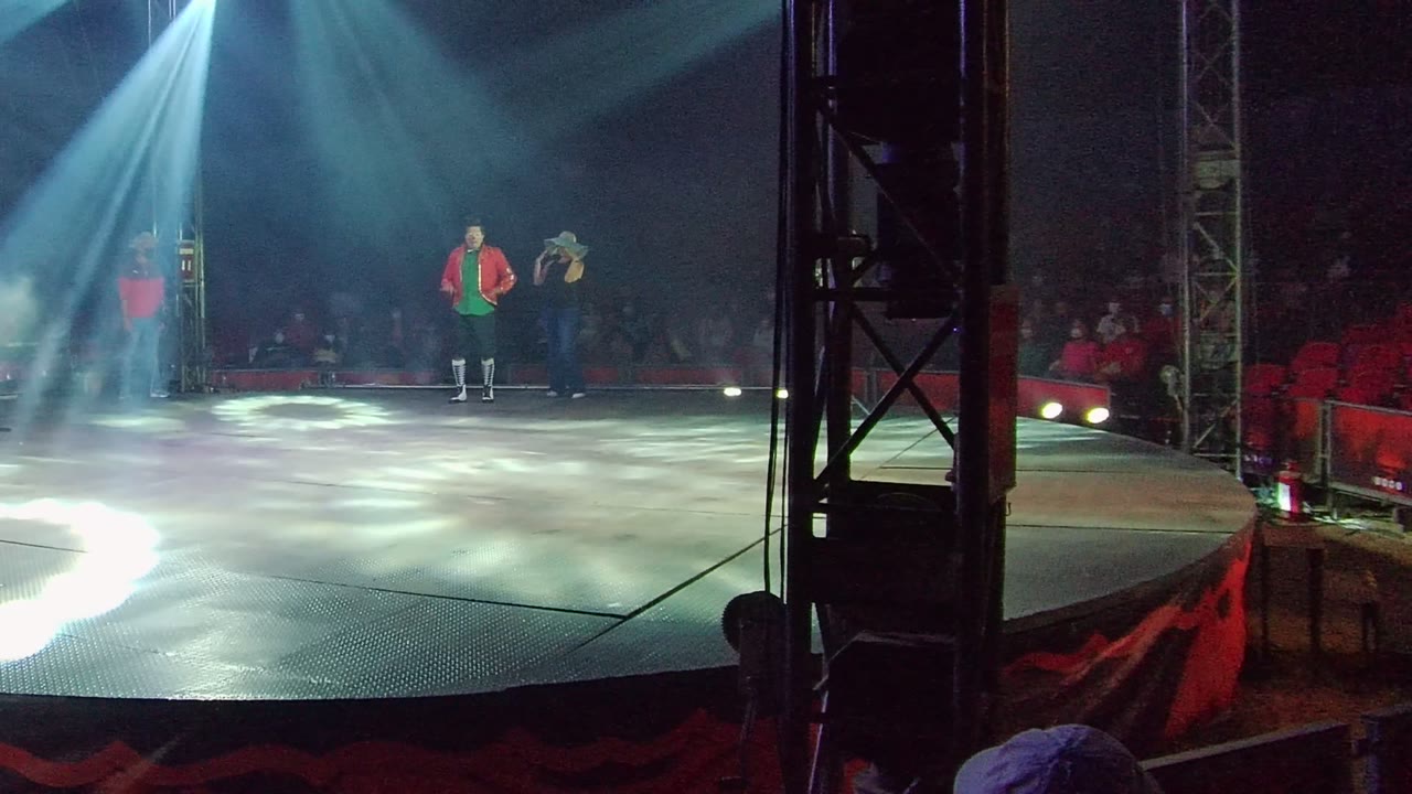 Two performers stand near center stage in San Jose del Cabo, with a third performer at left, pale spotlight beams overhead, and red seats visible behind them.