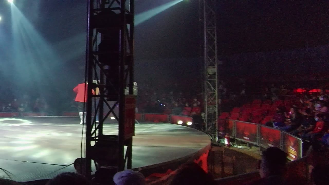 A red-shirted performer stands near the left side of a spotlighted stage in San Jose Del Cabo, Mexico, with a dark truss in the foreground and audience seating in the background.