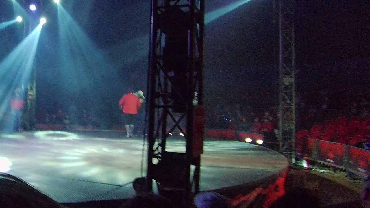 Red-shirted performer farther across the stage under blue-white beams.