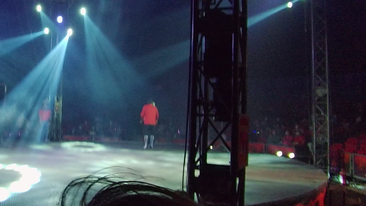 A red-shirted performer stands nearer the center of the stage in San Jose Del Cabo, Mexico, with red audience seating and overhead spotlights visible through the dark venue.