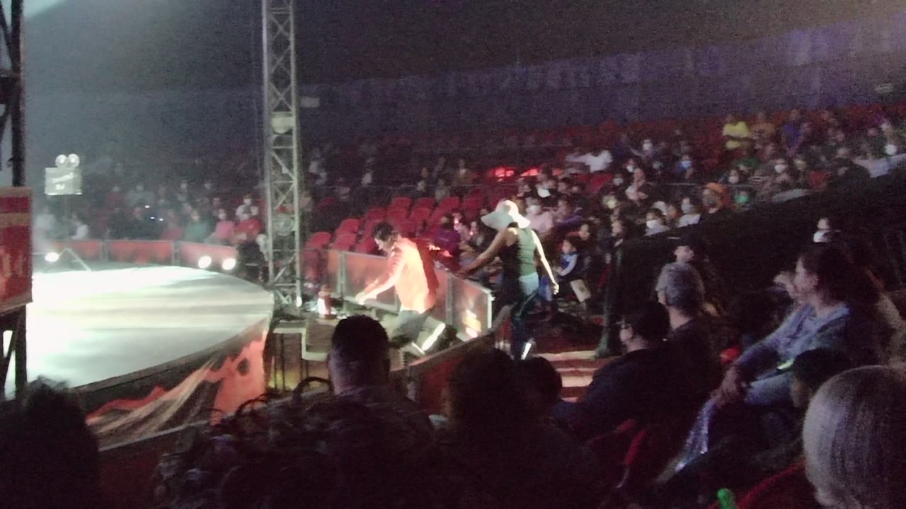 Two people stand near the edge of a lit indoor arena stage in San Jose del Cabo BCS Mexico while spectators sit in red chairs behind them.