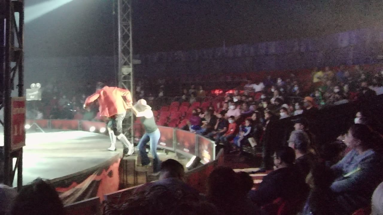The pair continues along the stage, one in a light hat and one in a bright top, with red arena seating and spectators filling the background.