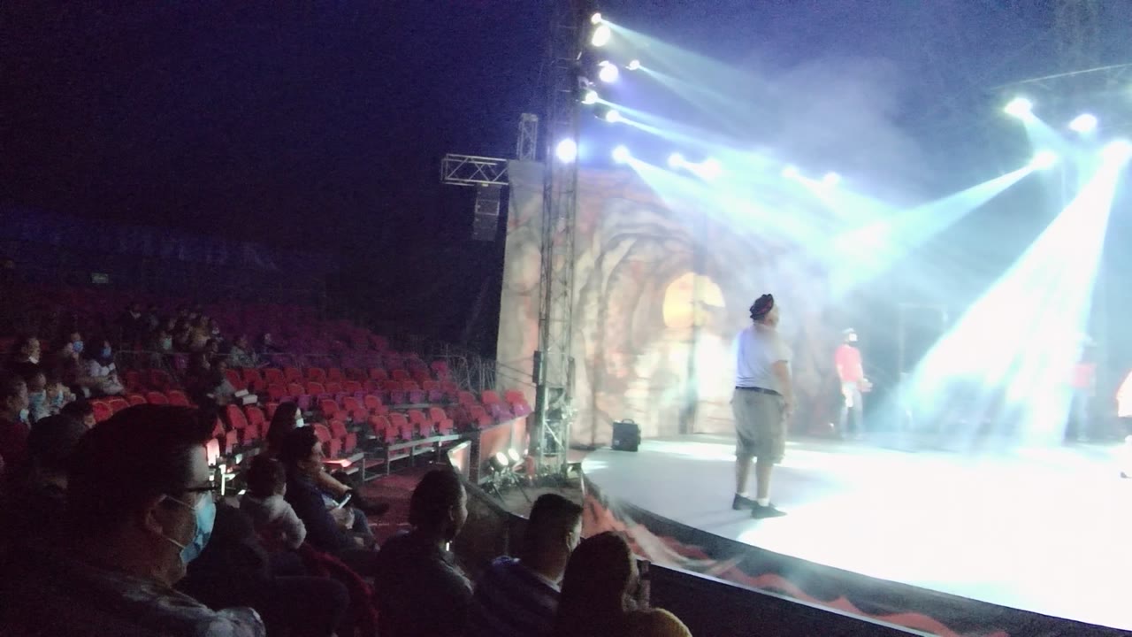In San Jose del Cabo, Mexico, audience members sit in red theater seats beside a bright stage where a performer stands under white spotlights and smoke.