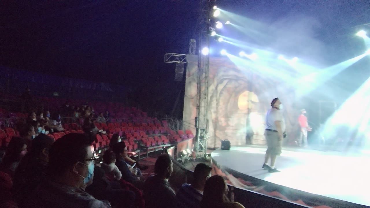 Audience in red seats watches a performer on a smoky stage