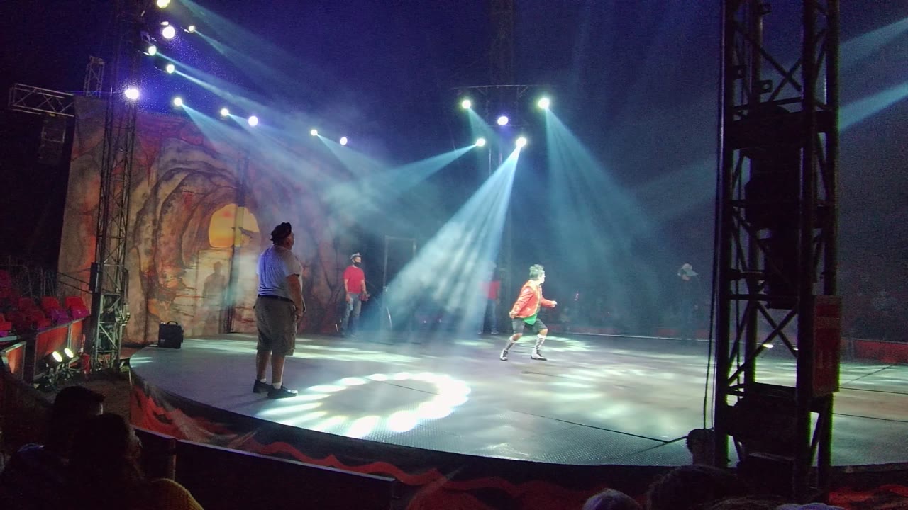 A San Jose del Cabo BCS Mexico stage performance begins with a dancer in orange moving across a lit circular stage, another performer in a blue shirt at left, and audience silhouettes in red seats.