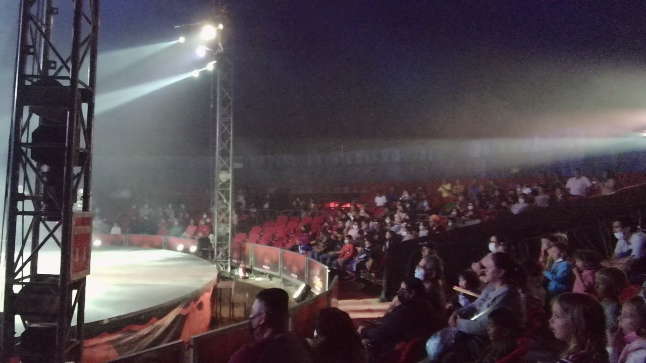 At 0:00 in San Jose del Cabo BCS Mexico, a circular circus stage is lit by white spotlights while rows of spectators sit in red seats beside a black truss.