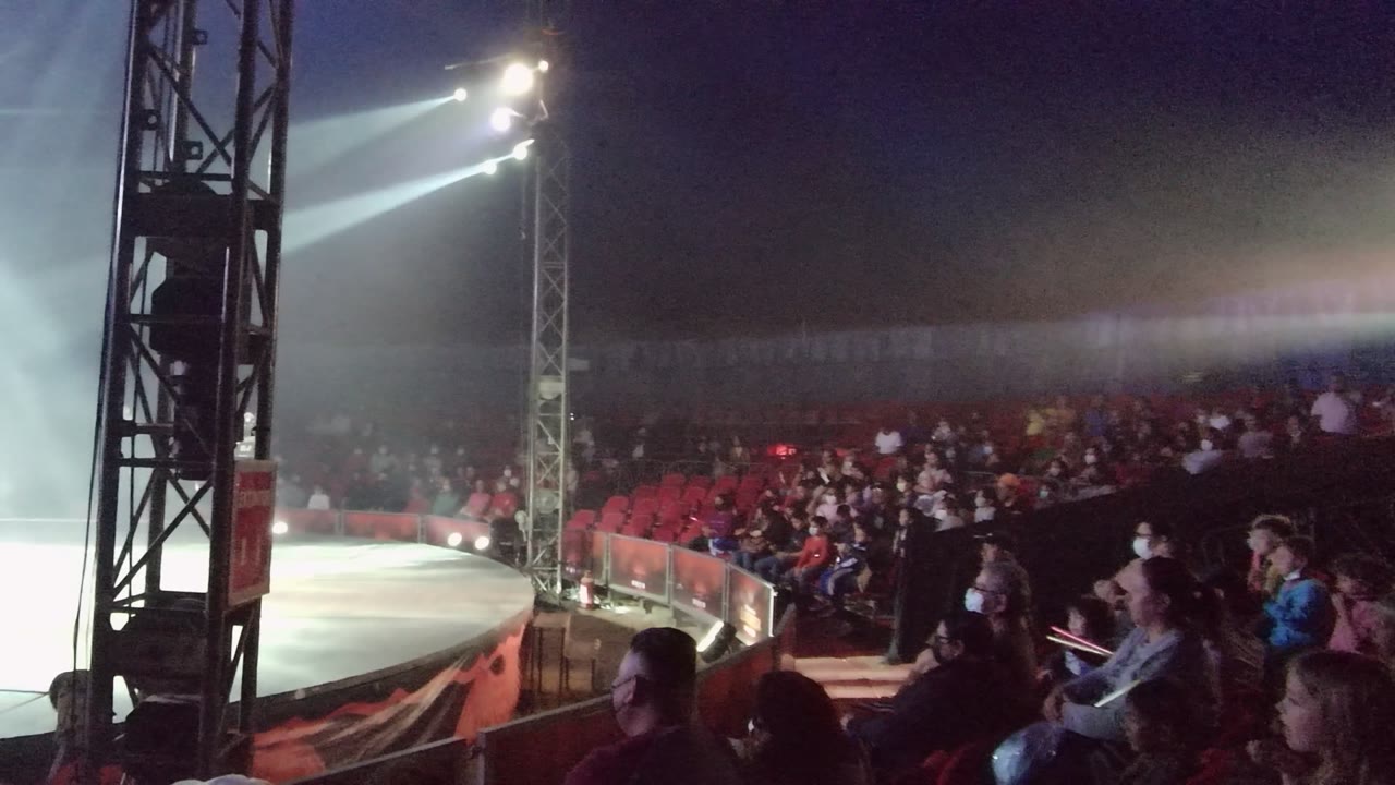 Audience watches a lit circus ring beside a stage truss