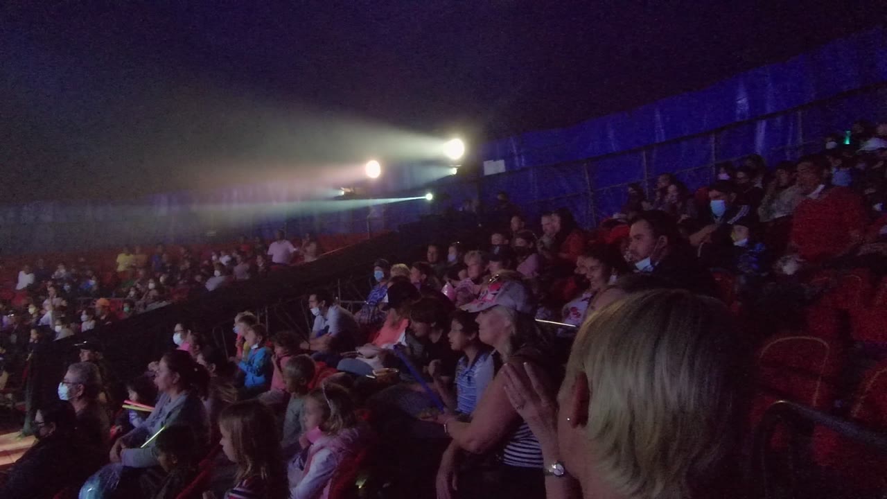 Audience members sit in red chairs inside a dark performance venue in San Jose Del Cabo BCS Mexico, with bright stage beams crossing the background and a blonde spectator close to camera.