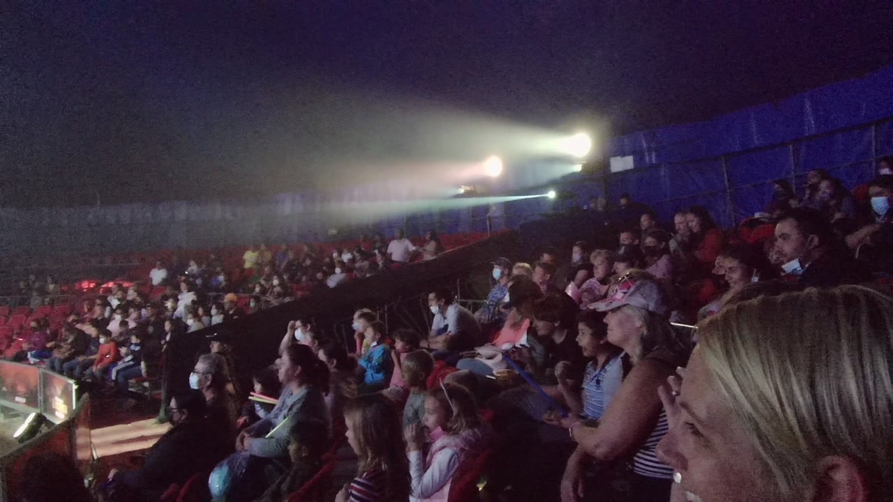 A side view shows seated spectators in San Jose Del Cabo watching toward the stage area, with masks visible on some faces and a bright light beam crossing the back.