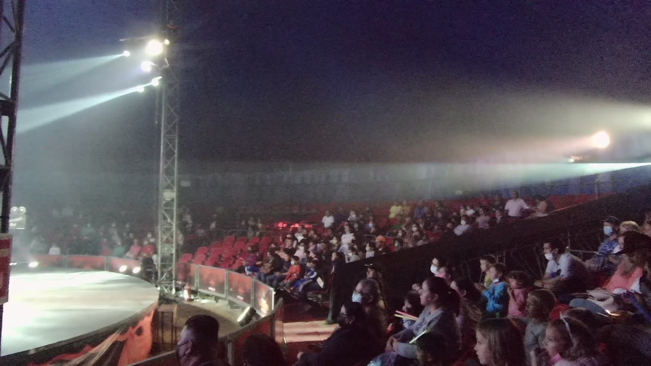 Crowd in red seats watching a stage area under bright beams.