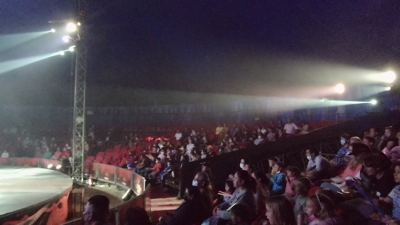 Spectators in San Jose Del Cabo BCS Mexico sit in rows near the stage, with several masked faces visible and strong white stage lights crossing above them.