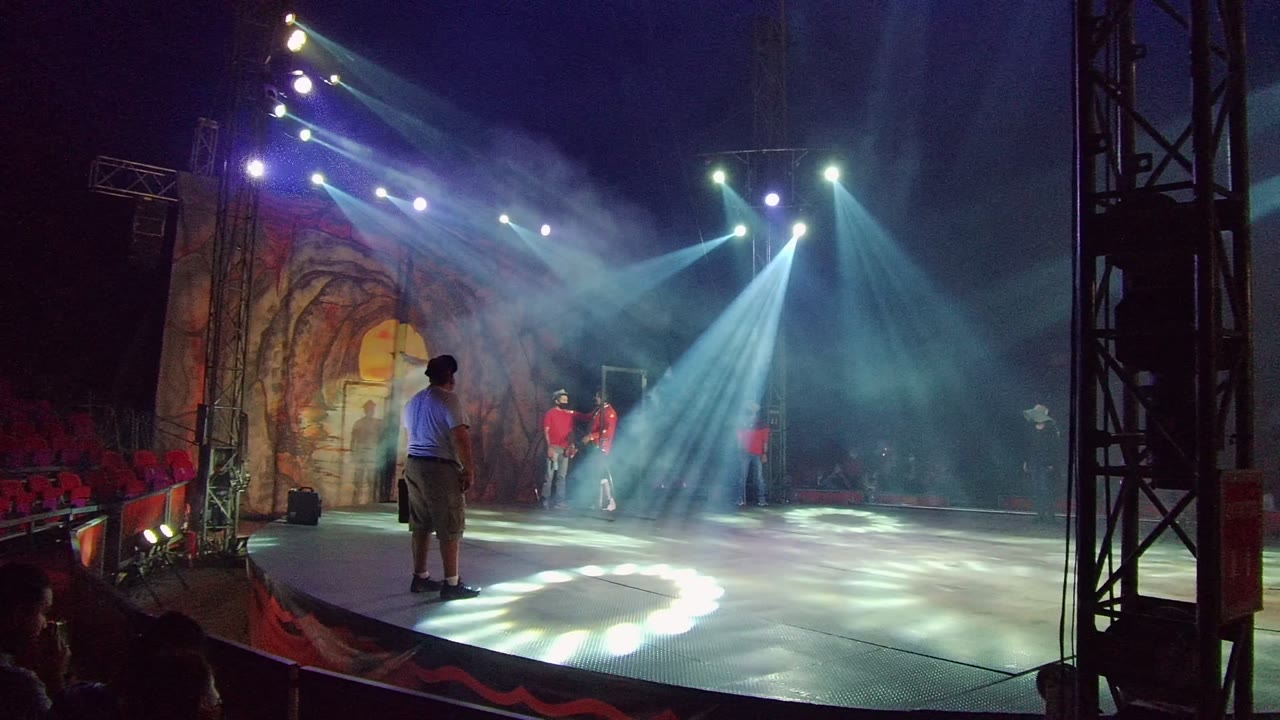 In San Jose del Cabo BCS Mexico, a person in a blue shirt stands near the front of a smoky stage as dancers in red shirts are lit by bright spotlights and audience seating appears behind them.