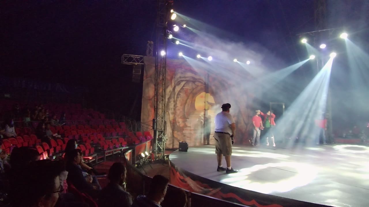 Wide view of a San Jose del Cabo stage performance with a foreground performer, dancers at the back, red audience seating, smoke, and crossing white-blue spotlights.