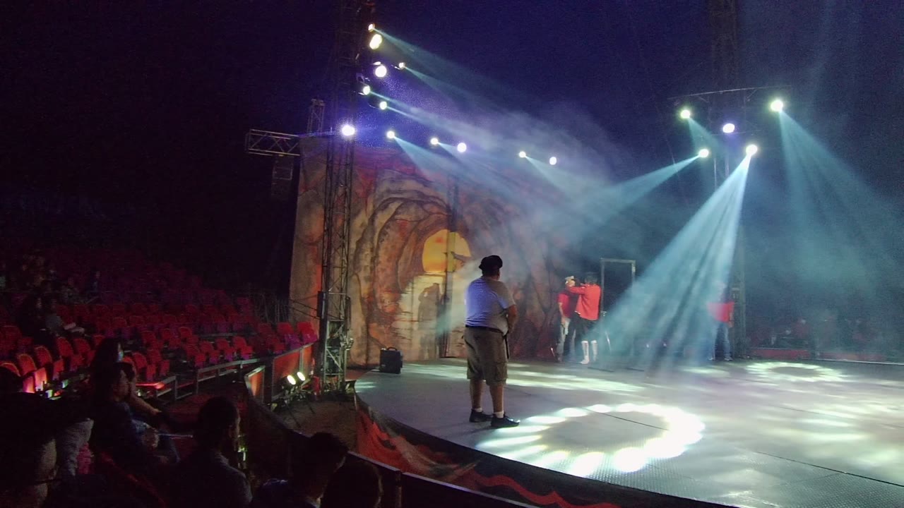 San Jose del Cabo performance frame with a red-clad dancer gesturing near the back of the stage, a foreground performer watching, red seats at left, and bright beams crossing the haze.