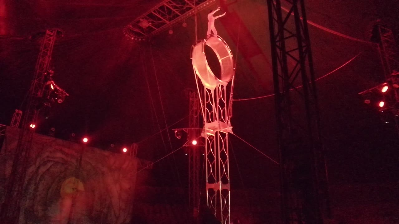 In San Jose del Cabo, Mexico, an aerialist stands atop a suspended circular ring inside a dark tent with red lights and rigging overhead.