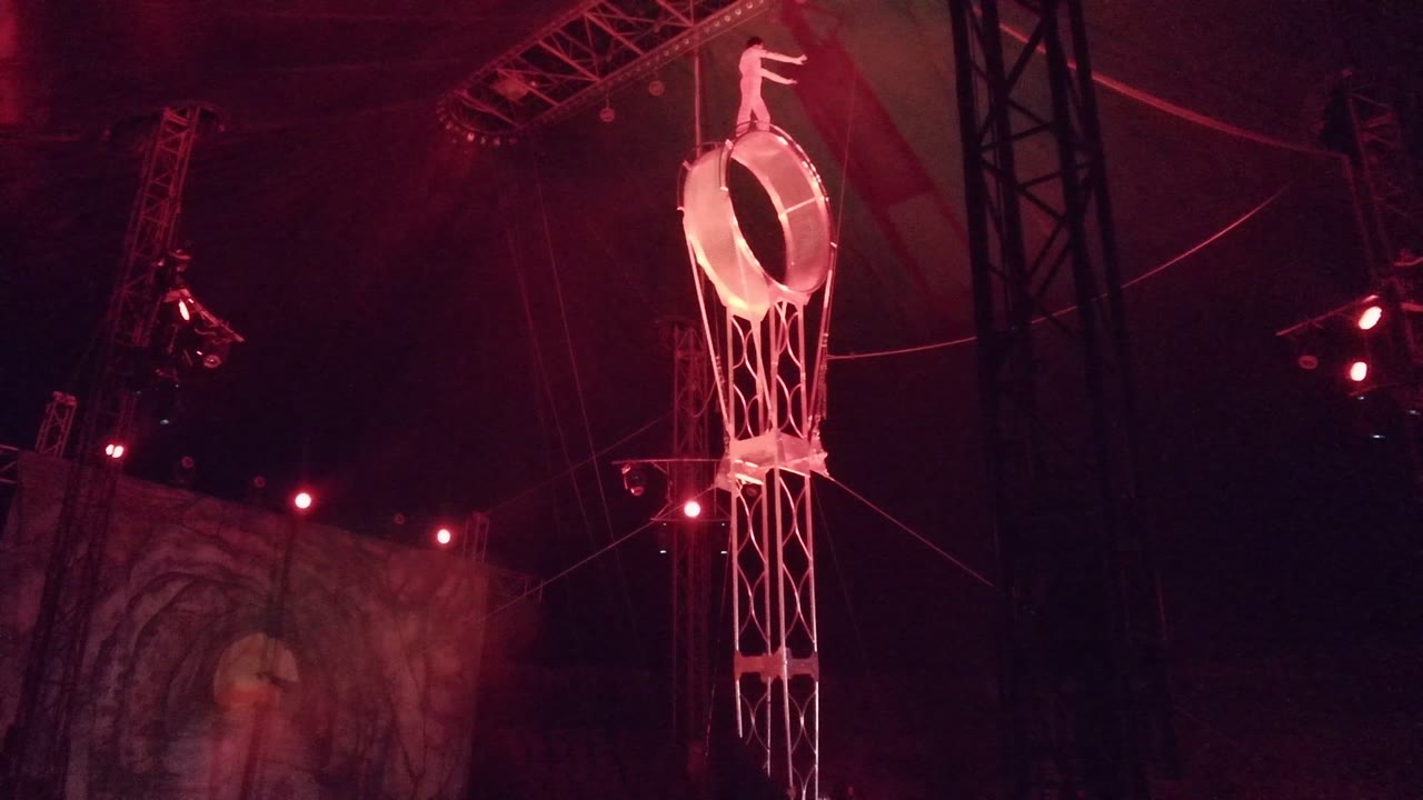 Aerialist posing above a suspended ring in a dark red-lit tent.