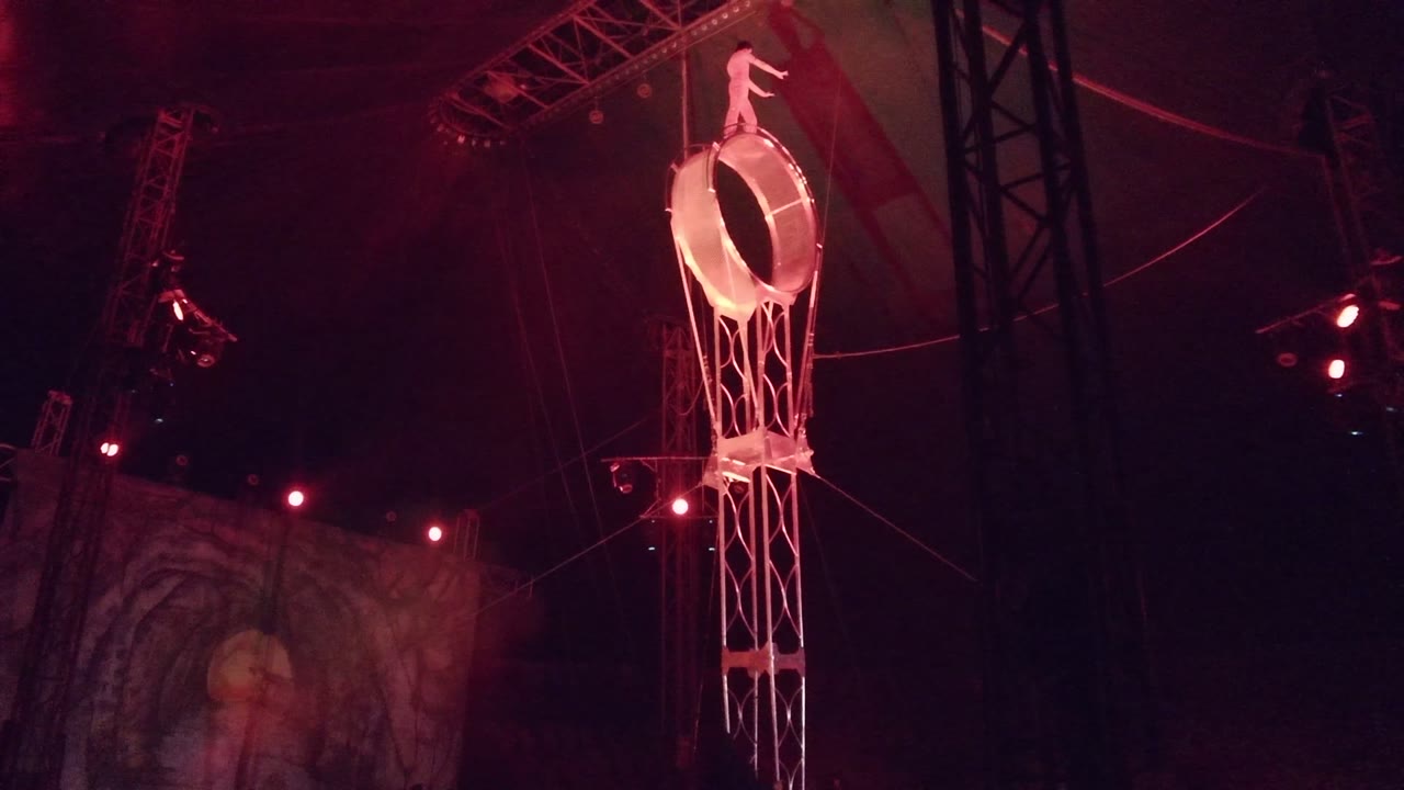 A red-lit circus ring hangs beneath tent truss in San Jose del Cabo, Mexico, with the aerialist posed above the circular frame.