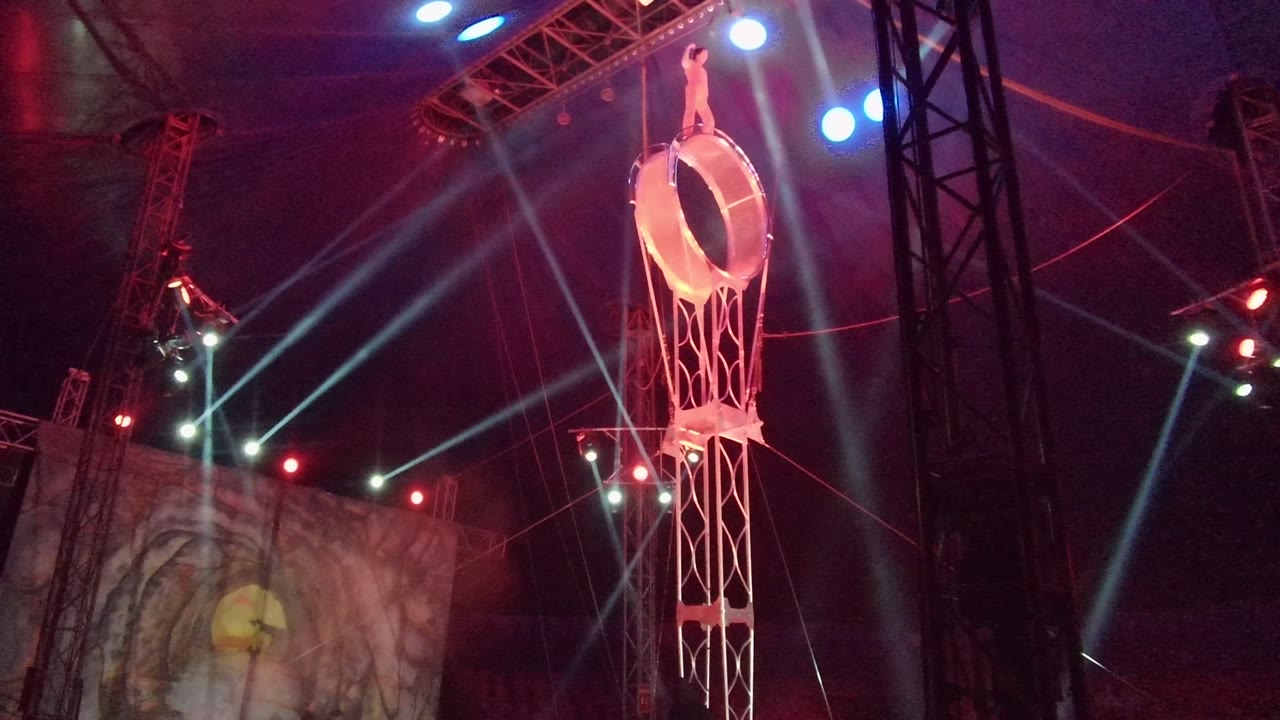 At 0:00 in San Jose Del Cabo BCS Mexico, a small aerial performer is high on a suspended circular ring above a tall truss, with red and blue stage lights and a face on the background screen.