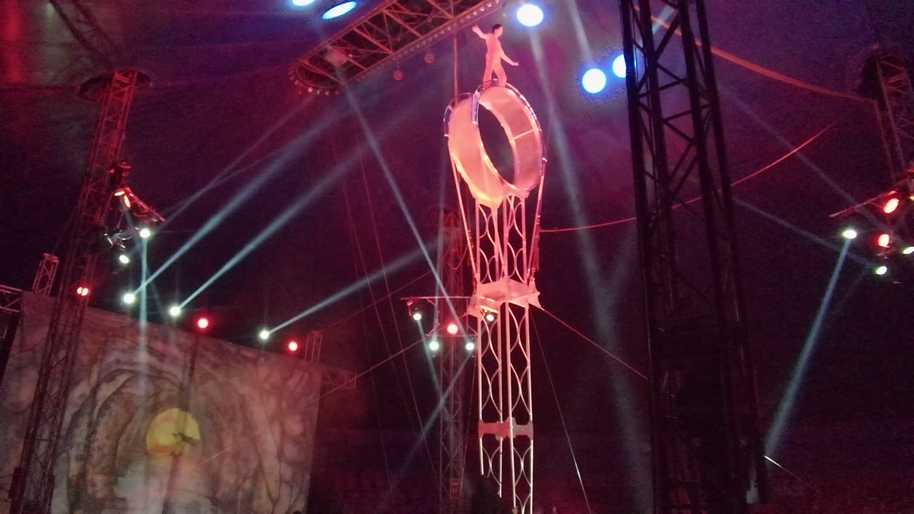 At 0:02 in San Jose Del Cabo BCS Mexico, the performer is still positioned on the suspended ring, with a bright blue spotlight near the apparatus and a face graphic visible on the stage screen below.