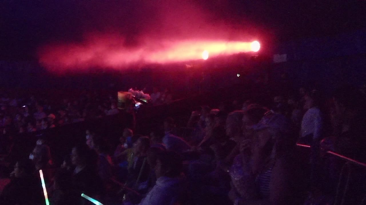 A San Jose del Cabo concert crowd sits and stands in darkness while red light burns through smoke above the audience.