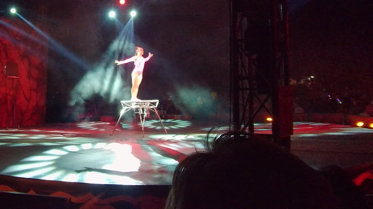 In San Jose del Cabo, Mexico, an acrobatic performer stands upright on a small stool while white overhead beams and red stage lighting frame the act.