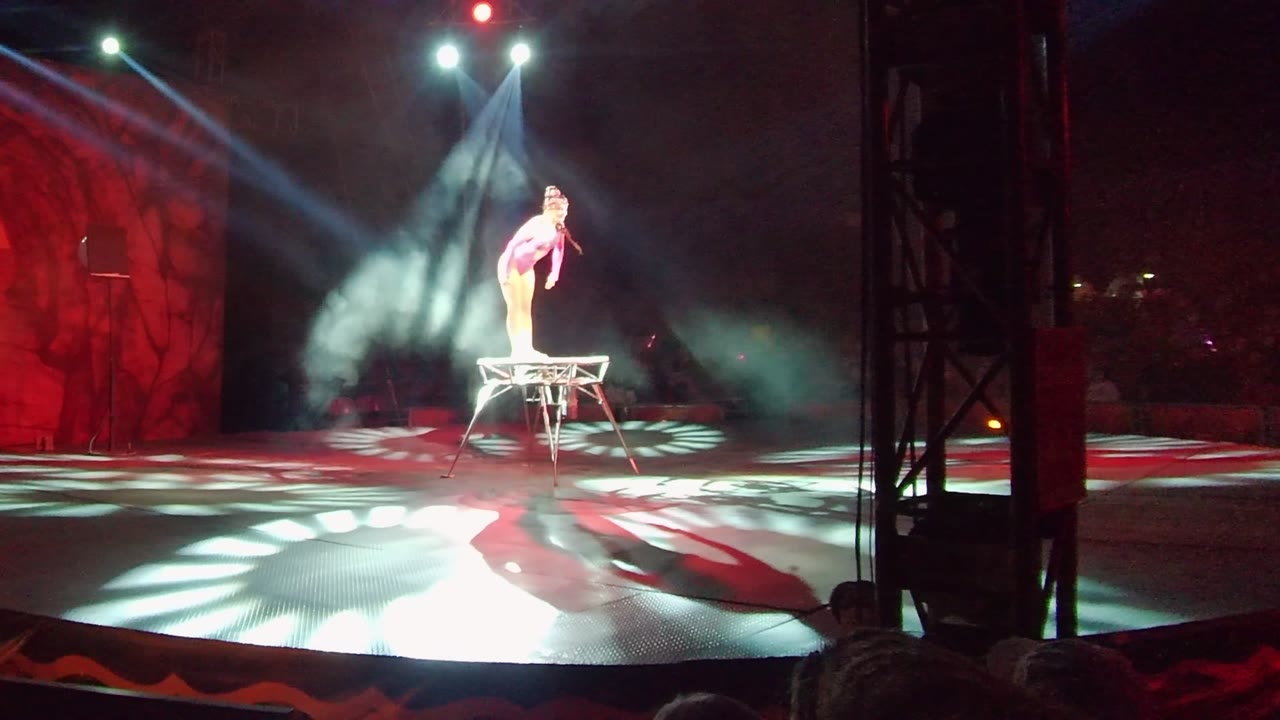 In San Jose del Cabo, Mexico, the performer holds a poised balance on the stool with one arm lowered, surrounded by red scenic light and white floor projections.