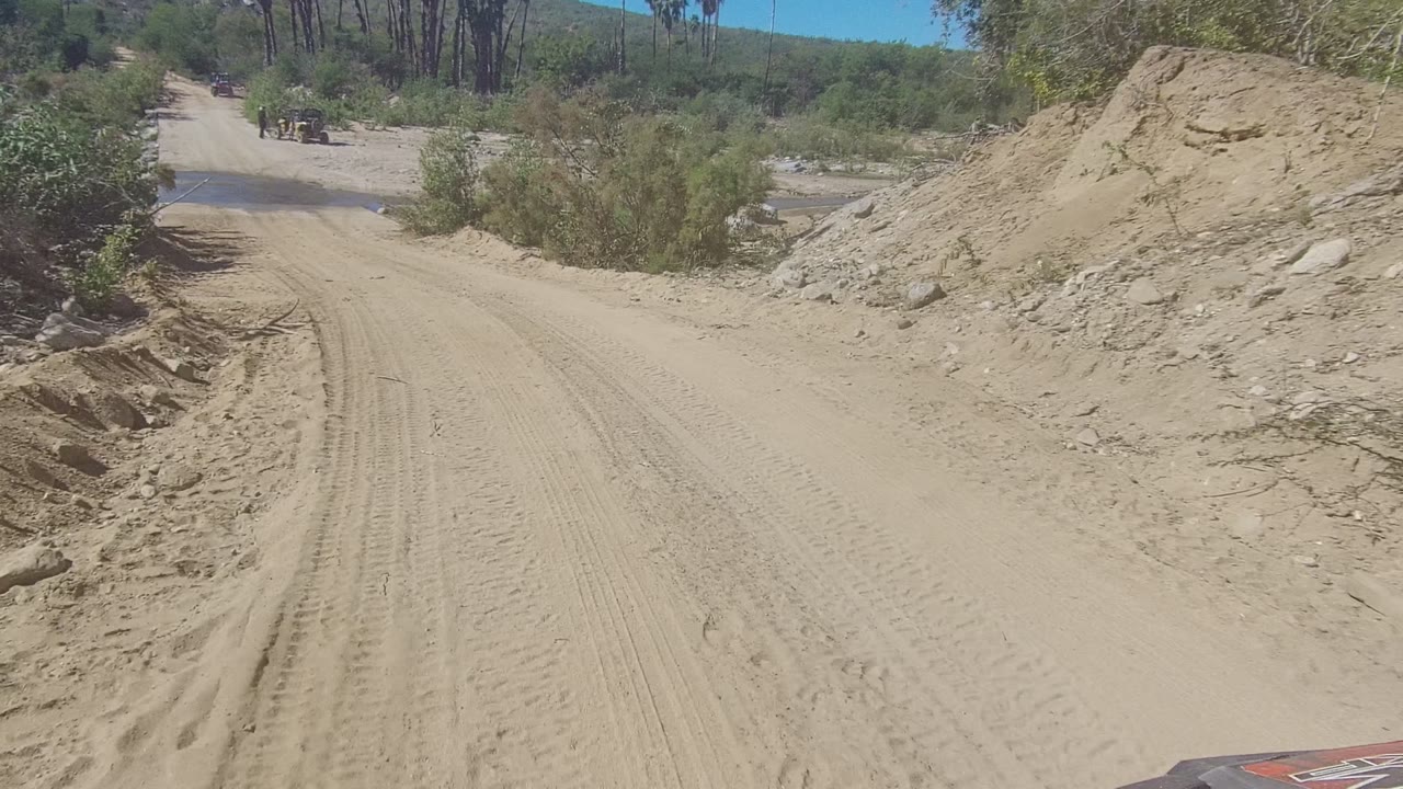 A forward road view in Los Cabos, Mexico shows the sandy track leading toward a darker water patch, with dry brush and rocks around it and no visible people.