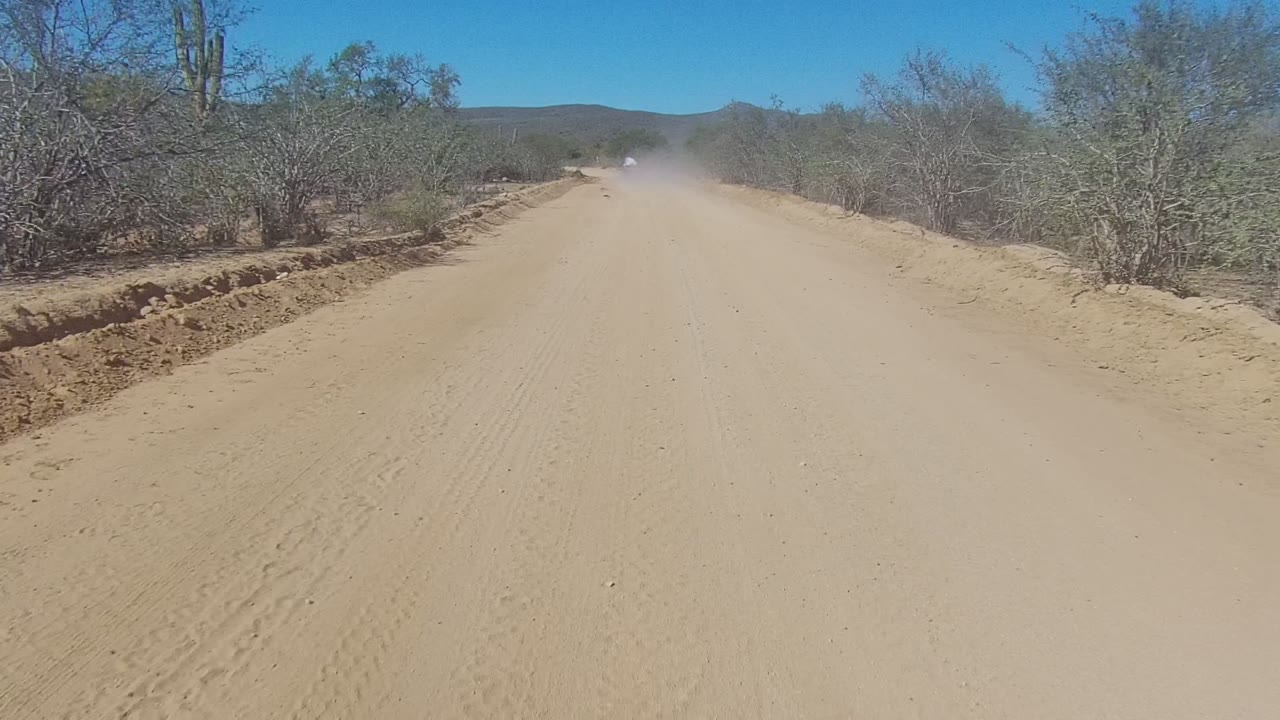At 0:00, a sandy road in Los Cabos BCS Mexico stretches toward a distant vehicle dust cloud, with dry shrubs, small trees, low hills, blue sky, and no visible people.