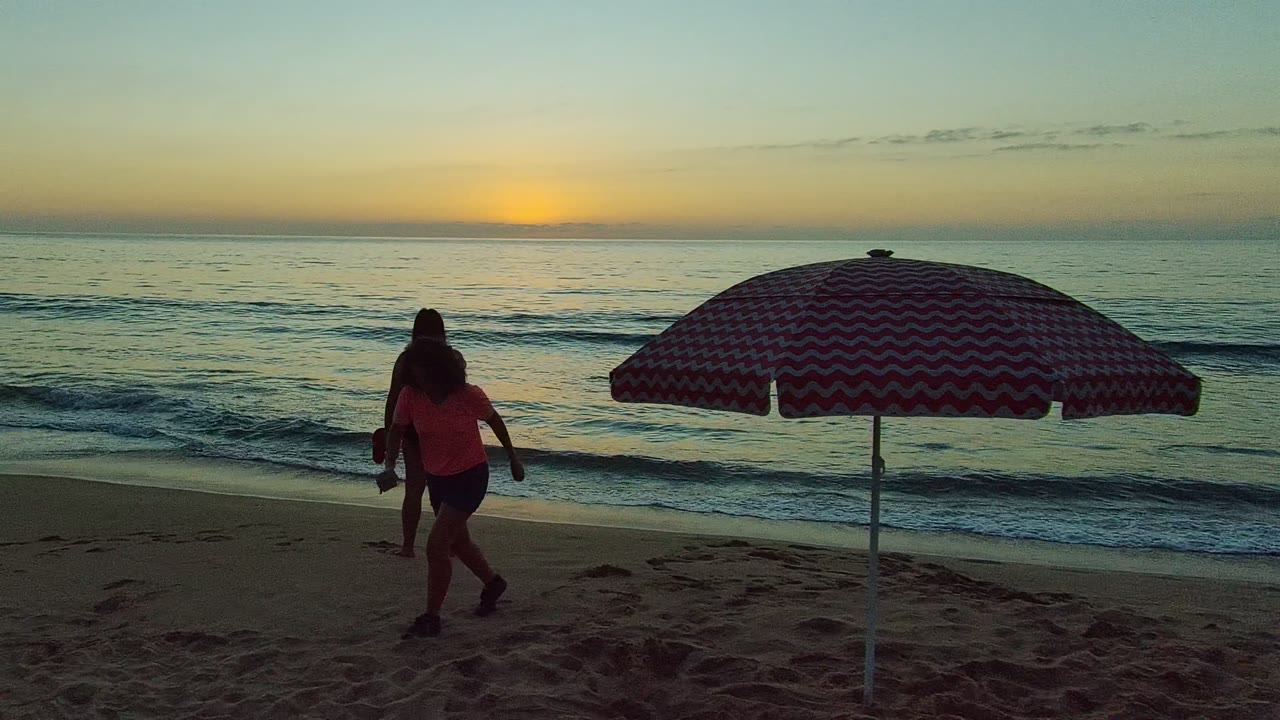 San Pancho Nayarit Mexico beach at sunset with a red and white striped umbrella in the foreground, one person walking at left, and another standing near the waterline.