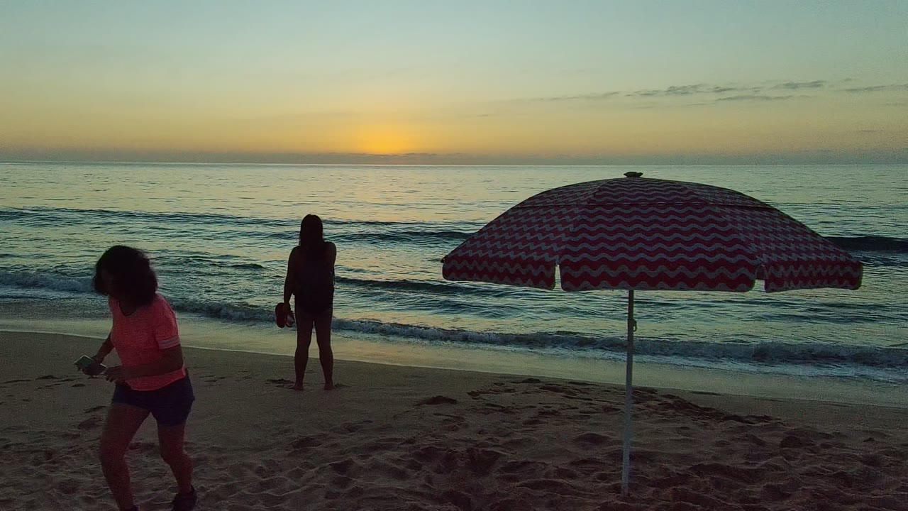 San Pancho Nayarit Mexico shoreline with the foreground person closer to camera under the edge of the striped umbrella and a second person standing near small waves.