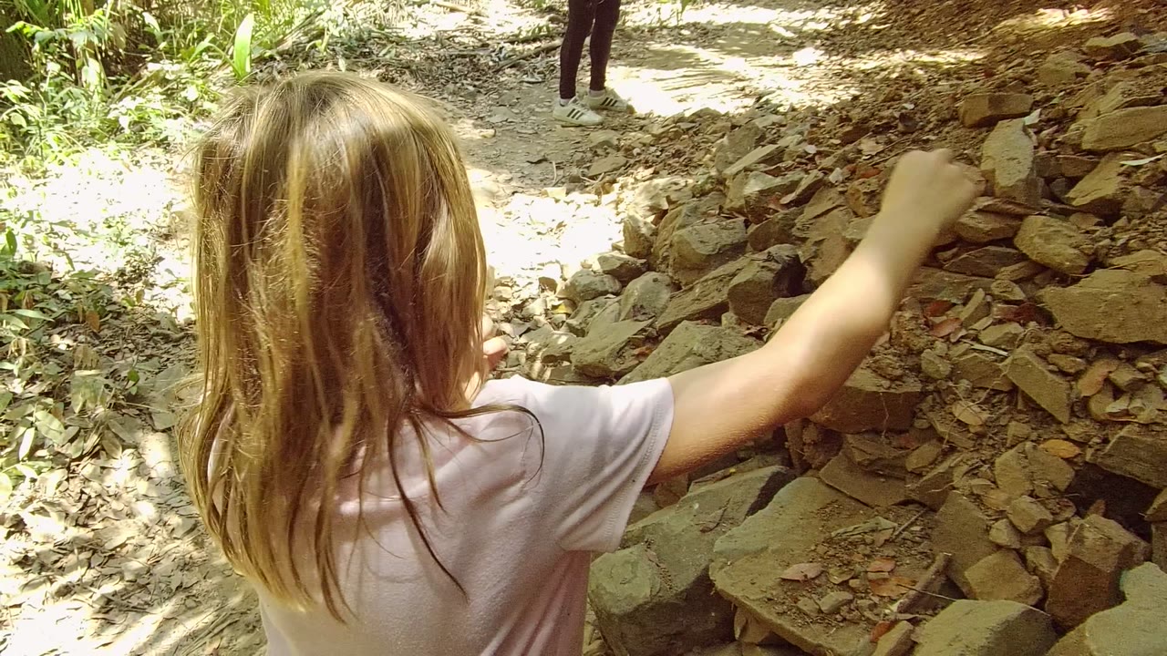 The girl is seen from behind in San Pancho Nayarit Mexico with one arm raised toward the rocky bank, while an adult stands farther along the bright dirt path.