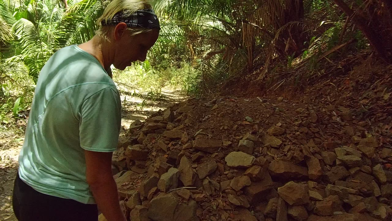 A woman stands at the left edge of a rocky dirt forest path in San Pancho Nayarit Mexico, with palm fronds and shaded vegetation around the trail.