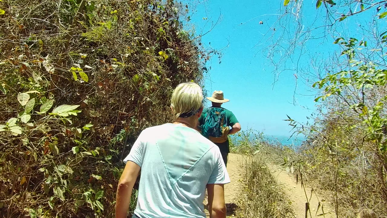 Two hikers walk away from the camera on a narrow dirt path in San Pancho Nayarit Mexico, with dry brush and green leaves framing a small glimpse of ocean ahead.