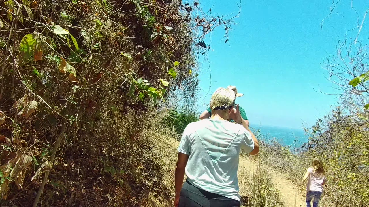 Two adult hikers are seen from behind on the San Pancho Nayarit Mexico trail while a child appears low at the right edge, with bright sky and ocean beyond the vegetation.