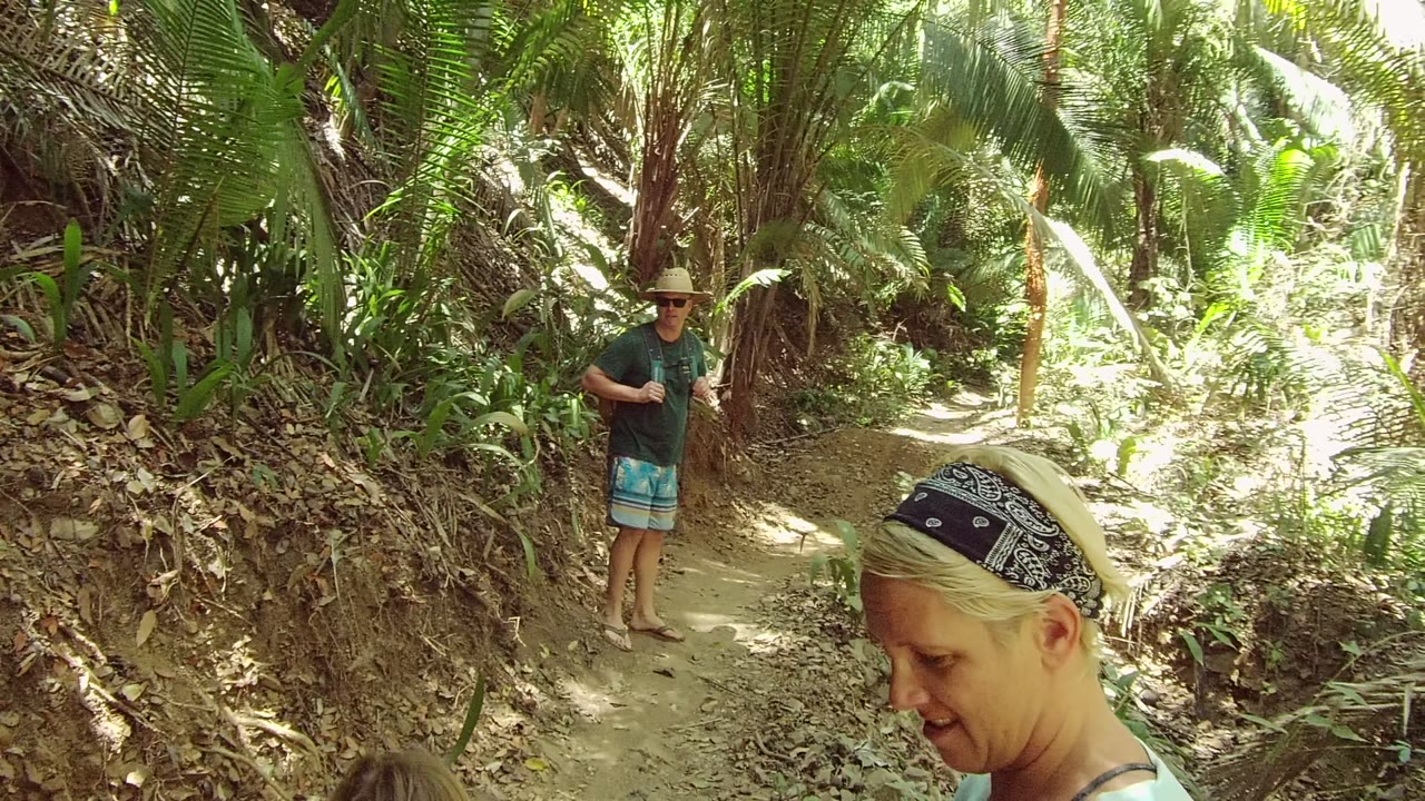 The woman remains close at right foreground while the man in a hat stands farther up the dirt path amid bright green forest growth in San Pancho Nayarit Mexico.
