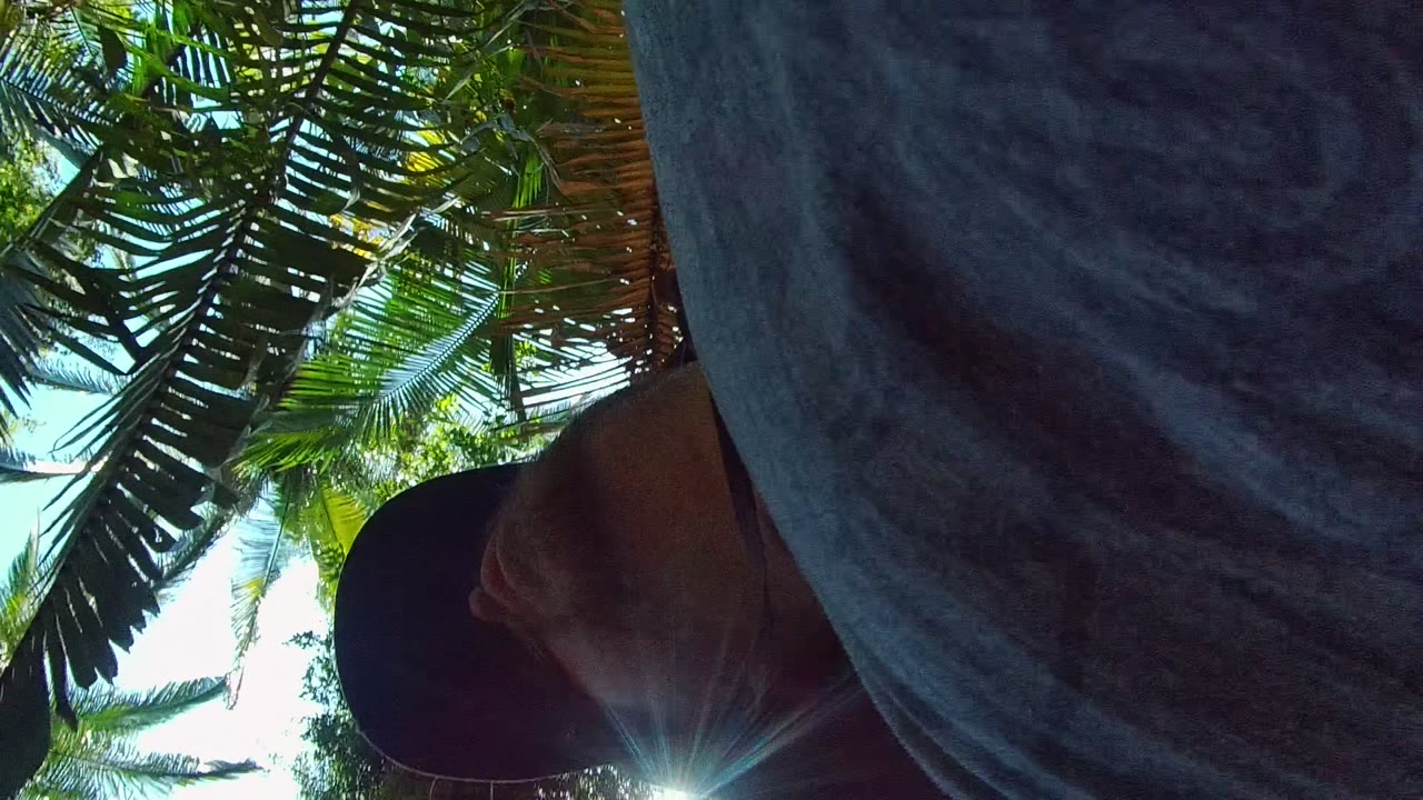 A close partial view of a person near the camera appears at the edge of the frame in San Pancho Nayarit Mexico, with dark clothing, a cap brim, sun flare, and dense leaves behind.