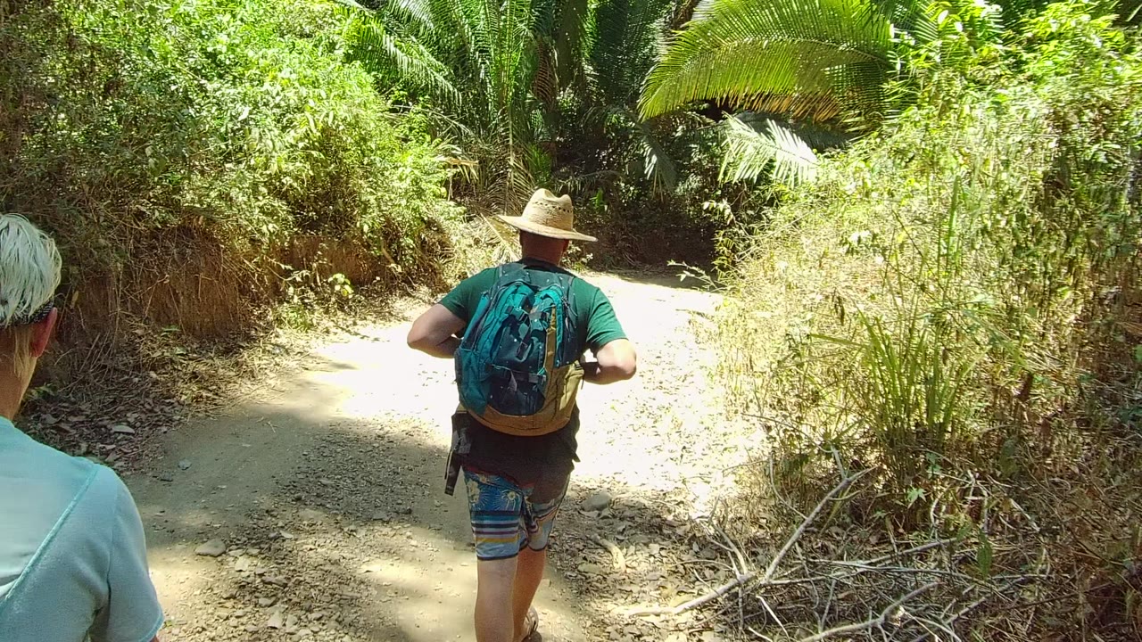 Two hikers walk away along a sunlit dirt trail in San Pancho Nayarit, Mexico, with a straw-hatted backpacker centered ahead and another person partly visible at left.
