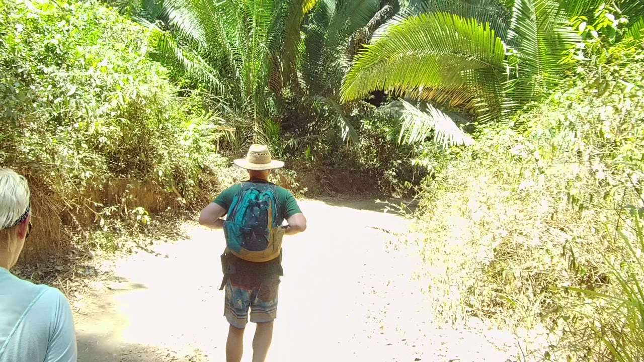 The backpacked hiker walks farther down the bright dirt trail in San Pancho Nayarit, Mexico, with the second hiker mostly out of frame at the left edge.