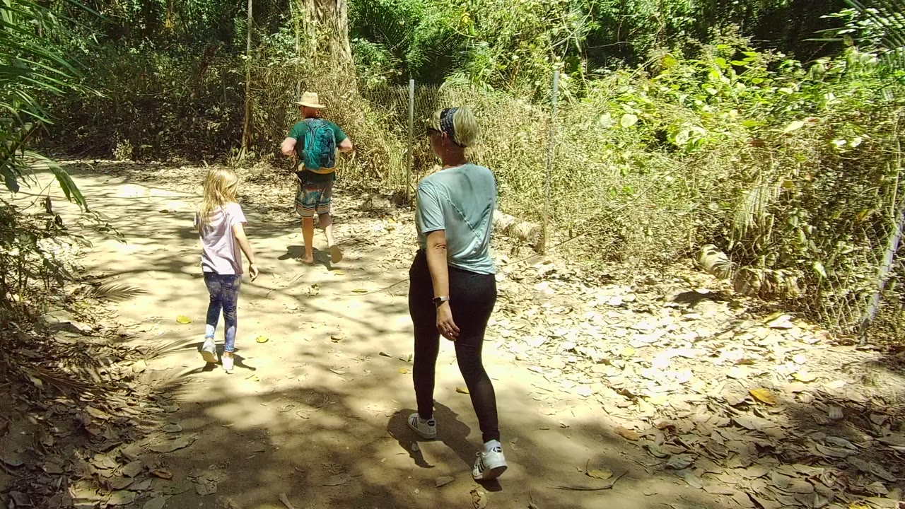 Three people walk away from camera on a dirt trail in San Pancho Nayarit Mexico, with the child at left, an adult ahead, and dense green foliage around the path.