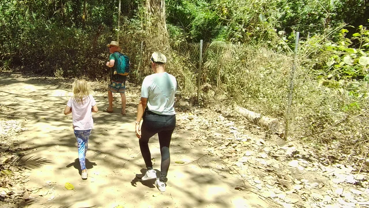 Three people seen from behind walk along the bright dirt path in San Pancho Nayarit Mexico, passing low dry brush and thick green vegetation under strong sunlight.